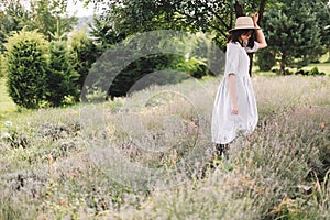 Stylish hipster girl in linen dress and hat relaxing in lavender field near tree. Happy bohemian woman enjoying summer vacation in