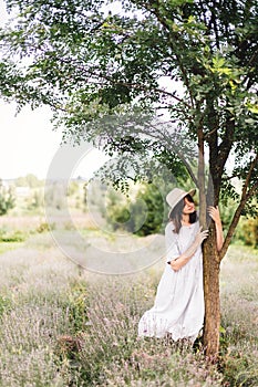 Stylish hipster girl in linen dress and hat relaxing in lavender field near tree. Happy bohemian woman enjoying summer vacation in