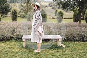 Stylish hipster girl in poncho, linen dress and hat posing at lavender field and relaxing in the morning. Bohemian woman enjoying