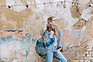 Stylish hipster girl in the retro jeans suit posing in front of the old brick wall. Trendy young woman with bag standing