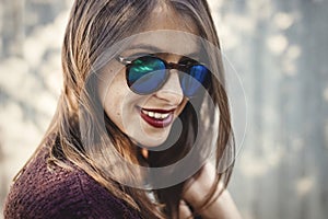 Stylish hipster girl smiling in sunny street on background of wooden wall. Boho girl in cool outfit and sunglasses posing in