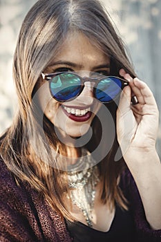 Stylish hipster girl smiling in sunny street on background of wooden wall. Portrait of boho girl in cool outfit and sunglasses