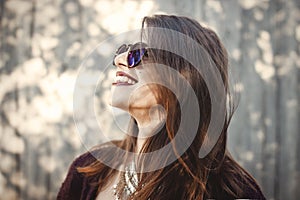 Stylish hipster girl smiling in sunny street on background of wooden wall. Portrait of boho girl in cool outfit and sunglasses