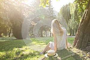 Stylish hipster girl with a straw hat is enjoying the sun outdoors. Happy woman sitting in blue dress near the tree. Travel