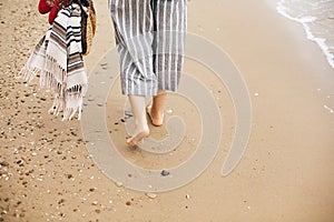 Stylish hipster girl walking barefoot on beach, holding bag and shoes in hand, closeup. Summer vacation. Space for text. Calm