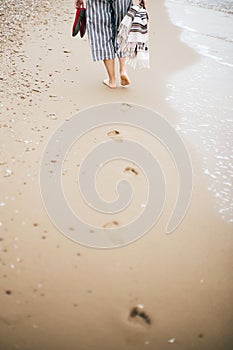 Stylish hipster girl walking barefoot on beach, holding bag and shoes in hand. Footprints on sand close up. Summer vacation. Space