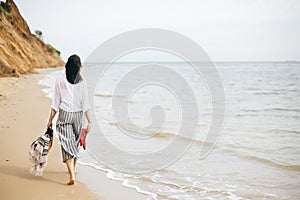 Stylish hipster girl walking barefoot on beach, holding bag and shoes in hand. Summer vacation. Space for text. Calm moment. Happy