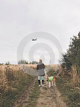 Stylish Hipster girl in yellow hat and coat walking with her golden dog and looking at plane in sky in autumn field. Woman in