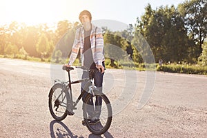 Stylish hipster guy standing near his bicycle while having rest after long trip, posing at camera, enjoying fresh air and beautifu