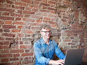 Young man freelancer working on his web site on portable laptop computer, sitting in modern interior against brick wall.