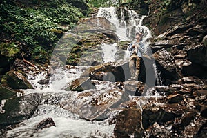 stylish hipster man in hat with photo camera, standing at waterfall in forest in mountains. traveler guy exploring woods. travel