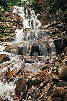 stylish hipster man in hat with photo camera, standing at waterfall in forest in mountains. traveler guy exploring woods. travel