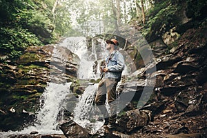 stylish hipster man in hat with photo camera, standing at waterfall in forest in mountains. traveler guy exploring woods. travel