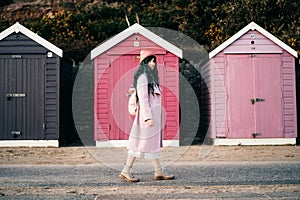 Stylish hipster woman with color hair in pink outfit and backpack walking along wooden beach huts on seaside. Off season