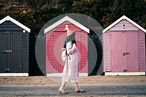 Stylish hipster woman with color hair in pink outfit and backpack walking along wooden beach huts on seaside. Off season