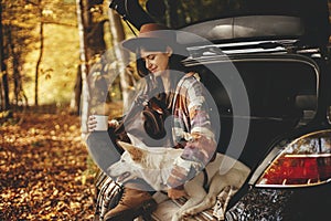 Stylish hipster woman with cup and backpack sitting with cute dog in car trunk in sunny autumn woods. Travel and road trip with