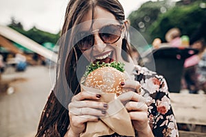 stylish hipster woman eating juicy burger. boho girl biting cheeseburger, smiling at street food festival. summertime. summer vac