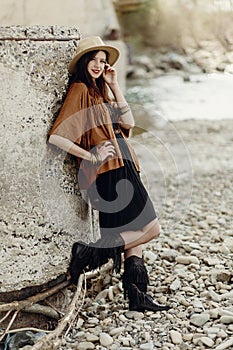 stylish hipster woman in hat and fringe poncho posing. boho traveler girl in gypsy look, near beach in mountains. summer travel.
