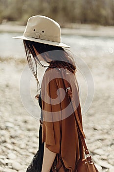 stylish hipster woman in hat, fringe poncho walking on river beach. boho traveler girl in gypsy look, summer travel. atmospheric