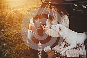 Stylish hipster woman in hat and sweater holding phone and sitting with cute dog in car trunk in sunset light. Travel and road