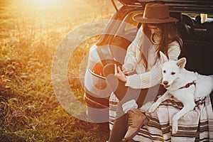 Stylish hipster woman in hat and sweater holding phone and sitting with cute dog in car trunk in sunset light. Travel and road