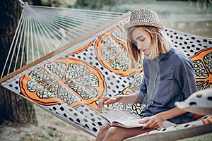 Stylish hipster woman reading book and relaxing in hammock in sunny summer park. blonde girl resting in forest, smiling and