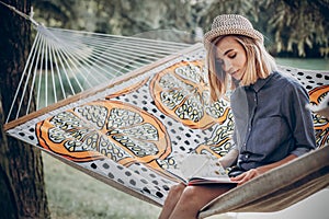 Stylish hipster woman reading book and relaxing in hammock in sunny summer park. blonde girl resting in forest, smiling and