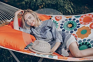 Stylish hipster woman relaxing in hammock in sunny summer park. blonde girl in fashionable hat resting in forest, smiling and