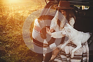 Stylish hipster woman in sweater holding phone and sitting with cute dog in car trunk in sunset light. Travel and autumn road trip