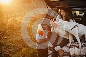 Stylish hipster woman in sweater holding phone and sitting with cute dog in car trunk in sunset light. Travel and autumn road trip