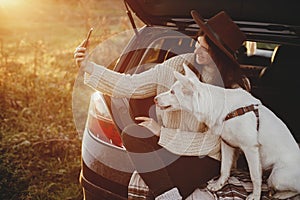 Stylish hipster woman taking selfie photo with cute dog in car trunk in warm sunset light. Autumn road trip with pet. Young female