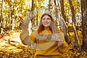 Stylish hipster woman traveler exploring map and showing path direction at autumn sunny forest