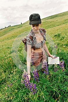 stylish hipster woman traveler holding in hand map and wildflowers and gathering lupin in mountains, travel and summer adventure