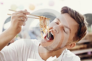 Stylish hungry man eating delicious wok noodles with vegetables from carton box with bamboo chopsticks. Asian Street food festival