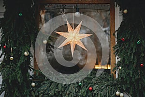 Stylish illuminated christmas star in window and fir branches with baubles on building exterior. Modern christmas decor in city