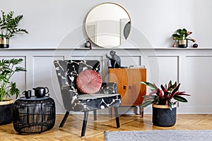 Stylish interior of living room with design armchair, wooden vintage commode, round mirror, shelf, plants, rattan coffee table.
