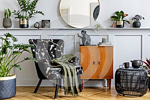 Stylish interior of living room with design armchair, wooden vintage commode, round mirror, shelf, plants, rattan coffee table.