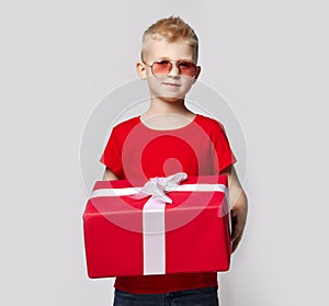 Stylish kid boy in red t-shirt and sunglasses stands holding big present for birthday gift box with ribbon in hands