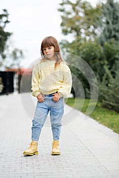 Stylish kid girl 5-6 year old wear trendy casual sweater and jeans pants walking in park outdoors.