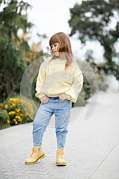 Stylish kid girl 5-6 year old wear trendy casual sweater and jeans pants walking in park outdoors.