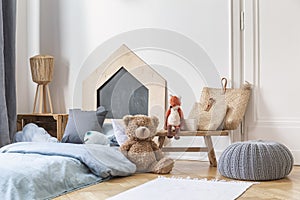 Stylish kid`s bedroom in tenement house. Grey pouf on the parquet, wooden table with bags and toys, bed with blue bedding