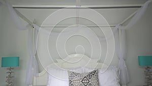 Stylish king-size bed with white baldachin, lot of white pillows, one is gray, two lamps on bedside tables on both sides