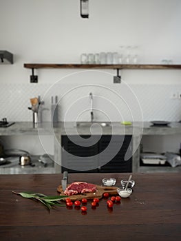 Stylish kitchen, on a wooden table there is a piece of fresh meat with spices, tomatoes and herbs, minimalist interior