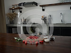 Stylish kitchen, on a wooden table there is a piece of fresh meat with spices, tomatoes and herbs, minimalist interior