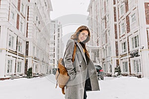 Stylish lady with brown backpack walking around city under snowfall. Outdoor photo of pretty girl with charming smile