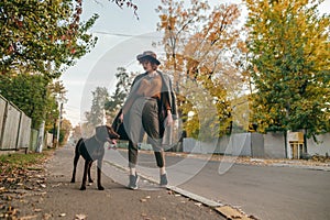 Stylish lady in vintage clothes posing at camera with dog on leash on the background of town street in autumn season. Curly lady