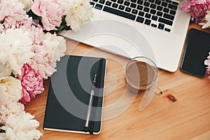 Stylish laptop, coffee cup, notebook, pen, pink and white peonies on wooden table with space for text. Freelance concept.