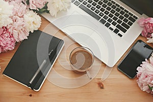 Stylish laptop, phone with empty screen, black notebook, coffee and peonies on rustic wooden table. Freelance concept. Girly