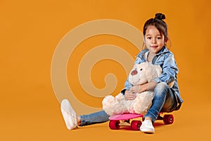 Stylish little child girl with skateboard in denim on orange background