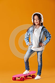 Stylish little child girl with skateboard in denim on orange background